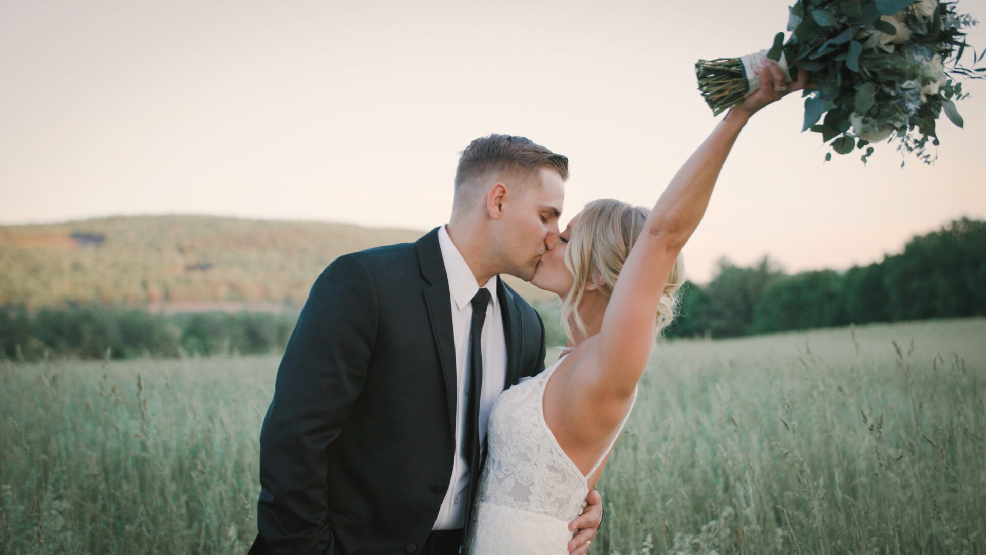 Wedding couple kissing in field for video. Bride holding bouquet over her head