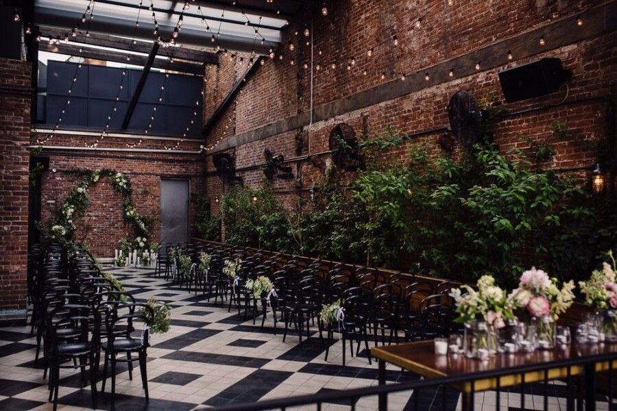 Industrial style wedding venue with brick walls, green trees, string lighting and black chairs decorated with flowers