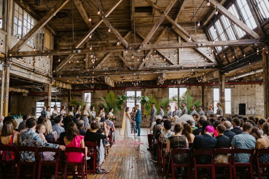 Bride and Groom getting married in rustic wooden venue with string lights in front of guests