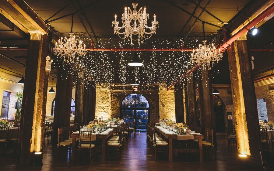 Wedding reception venue with string lights, lit chandeliers and wooden tables decorated with flowers and candles