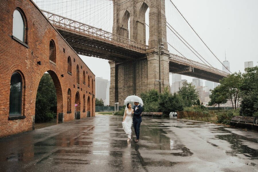 Bride and Groom walking in rain while holding clear umbrella under bridge